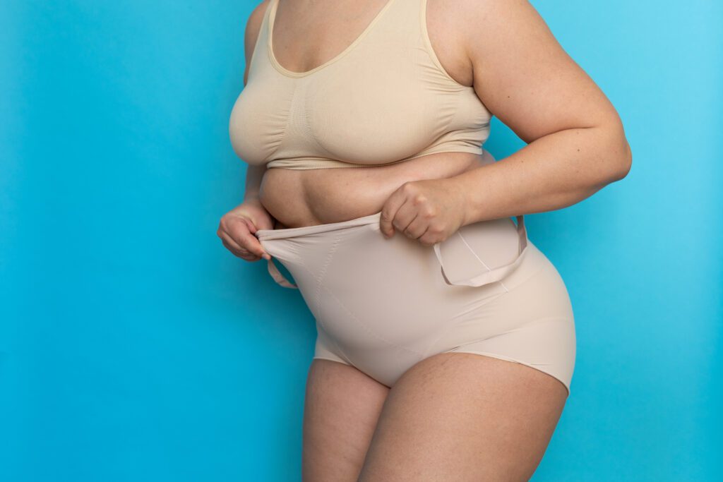 Plus size woman in beige underclothes pulling shapewear panties over belly closeup. Tighten figure and wear lifting underwear for weight loss, blue background. Concept of diet and fighting overweight.